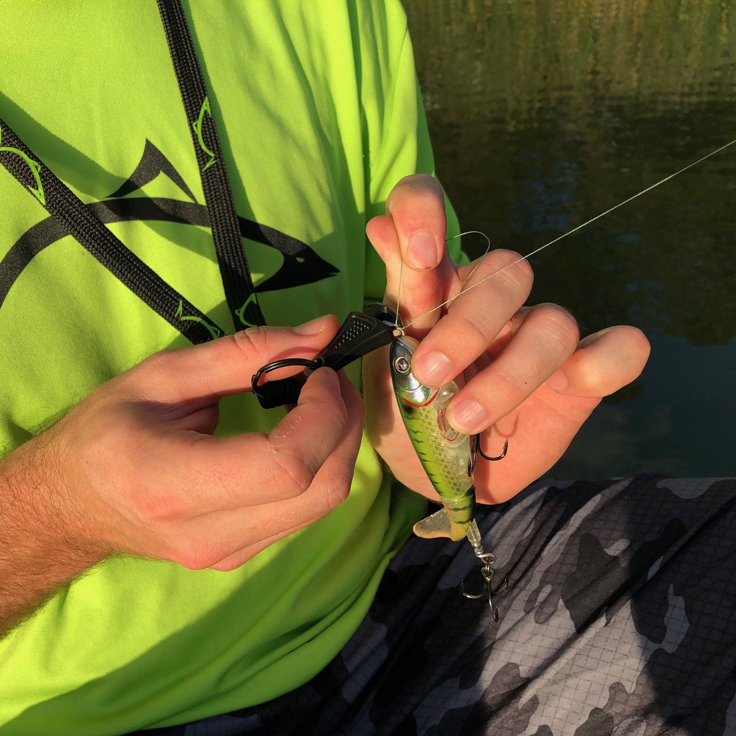 Wearing a bright green shirt, a person near water uses pliers and the Line Cutterz "TRIPLE PLAY" Fishing Line Cutter Multi-Pack to attach a small fish-shaped lure to their fishing line.