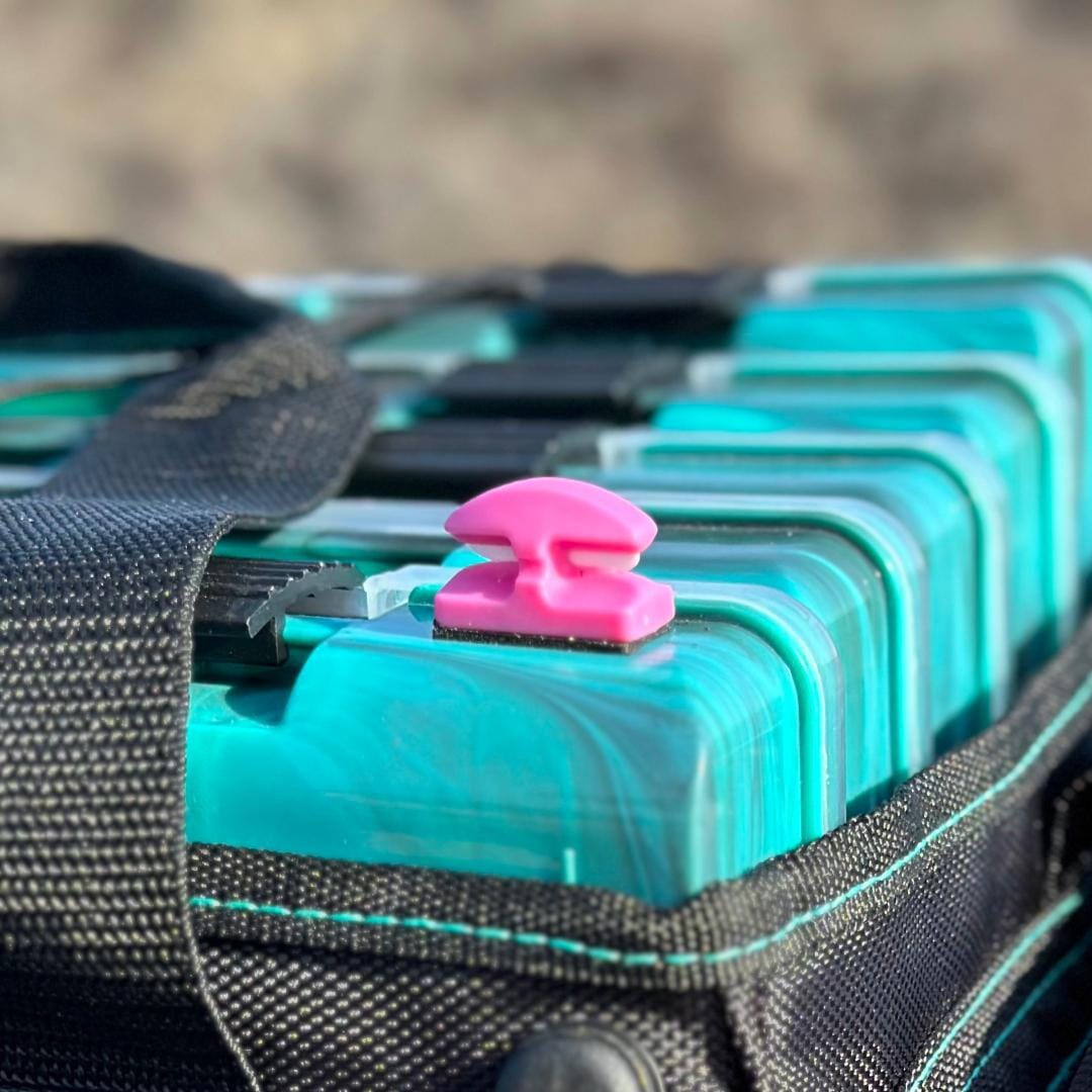 A close-up of a teal tackle box with a black zipper and a pink Line Cutterz Ceramic Blade Peel & Stick Flat Mount line cutter on top, outdoors with a blurred natural background.