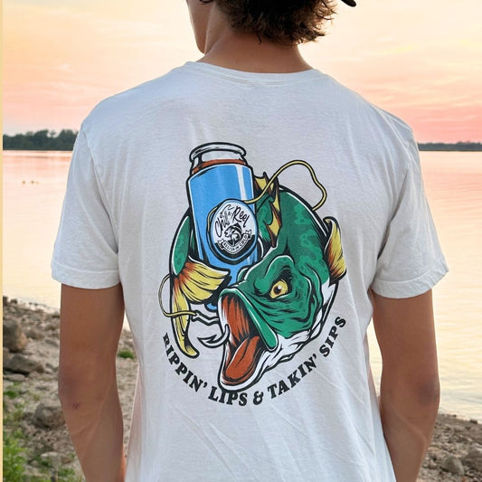 A person with long hair wearing a super soft cotton white Chill-N-Reel Soft Short Sleeve T-shirt - Rippin Sippin FIGHTER FISH with Chill-N-Reel featuring an illustrated green fish holding a can. The text on the shirt reads, "Rippin' Lips & Takin' Sips." The person is standing near a body of water with a serene pink and orange sky in the background.