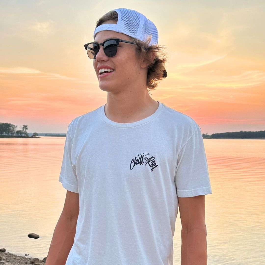 A young person wearing sunglasses, a white cap, and a Soft Short Sleeve T-shirt - Rippin Sippin FIGHTER FISH with Chill-N-Reel emblem stands by a calm lake during a colorful sunset. The sky is shades of orange and pink, reflecting on the water. The person appears to be smiling.