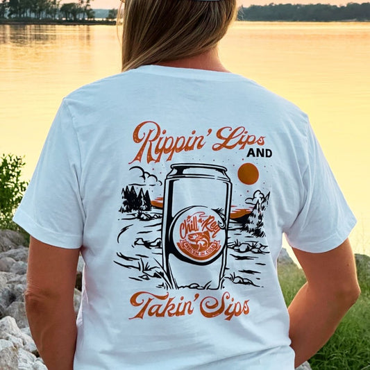 A person stands near a large body of water at sunset, wearing a white T-shirt. The shirt features a groovy mountain landscape design with the text "Soft Short Sleeve T-shirt - Rippin Sippin LANDSCAPE with Chill-N-Reel" and "Chill-N-Reel." Trees and a serene shoreline are visible in the background, enhancing the tranquil scene.