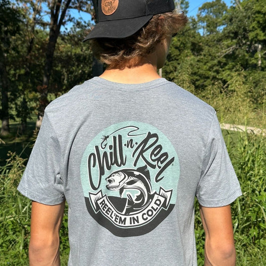 Person standing outdoors with their back to the camera, wearing a super soft cotton gray Soft Short Sleeve T-shirt - Chill-N-Reel CLASSIC COLOR LOGO (on BACK) and a dark cap. The T-shirt features a logo with a leaping fish and the words "Chill-N-Reel, Reel Em In Cold" prominently displayed. The background is lush with greenery.