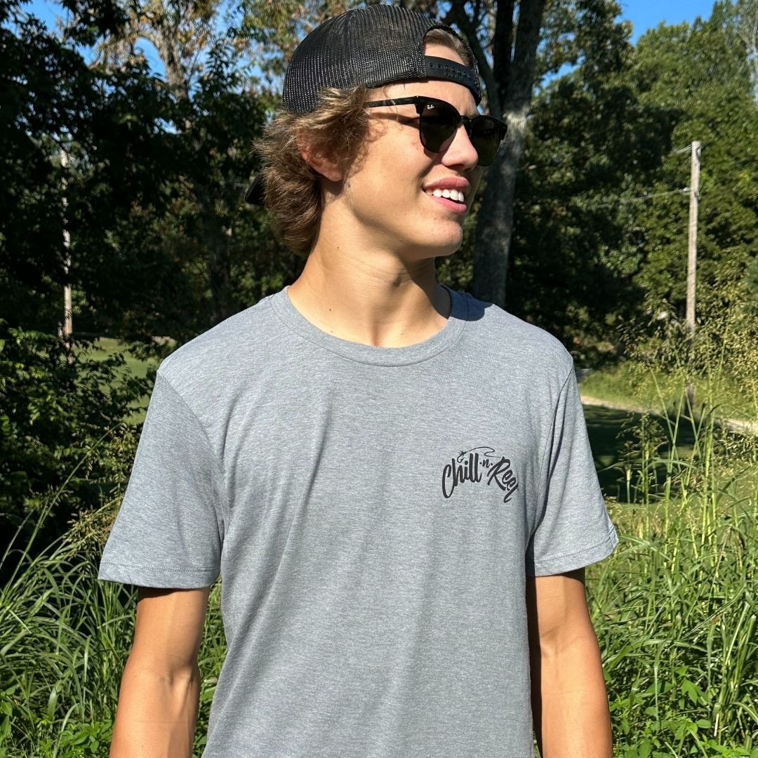 A young man with light brown hair is smiling while standing outdoors. He is wearing a backward baseball cap, dark sunglasses, and a light gray t-shirt made of super soft cotton with the text "Chill-N-Reel" on the chest. Behind him are trees and tall grass under a clear sky. The t-shirt he is wearing is the Soft Short Sleeve T-shirt - Chill-N-Reel CLASSIC COLOR LOGO from Chill-N-Reel.
