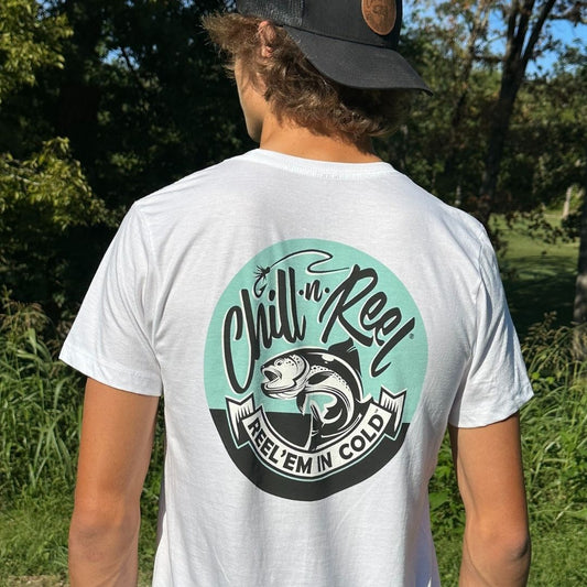 A person wearing a Chill-N-Reel Soft Short Sleeve T-shirt - Chill-N-Reel CLASSIC COLOR LOGO (on BACK) made of super soft cotton with a circular design on the back that features a fish and the text "Chill-N-Reel" and "Reel 'Em In Cold." The individual is standing outdoors with greenery and trees in the background, and wearing a black hat.