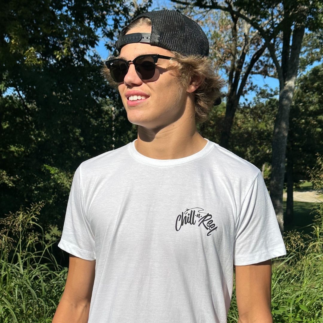 A young man stands outdoors wearing a Chill-N-Reel Soft Short Sleeve T-shirt - Chill-N-Reel CLASSIC COLOR LOGO (on BACK). He has on dark sunglasses and a backward black cap. The background features greenery and trees under a clear blue sky, perfectly complementing his Chill-N-Reel vibe.