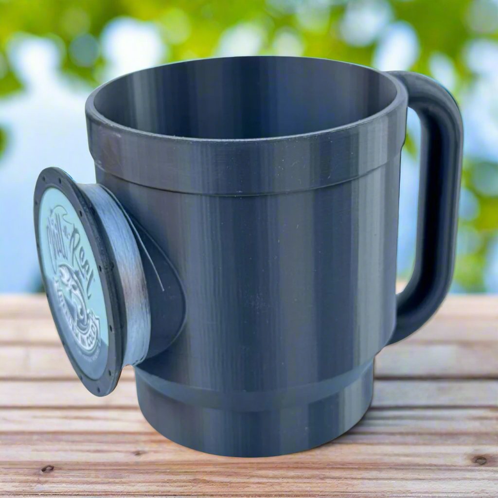 A black 30oz tumbler equipped with a Chill-N-Reel® Tumbler Handle sits on a wooden surface, showcasing a fish logo alongside the text "Reel Cool Dad." In the background, blurred green foliage and a touch of blue sky create an ideal setting for any fishing game enthusiast.