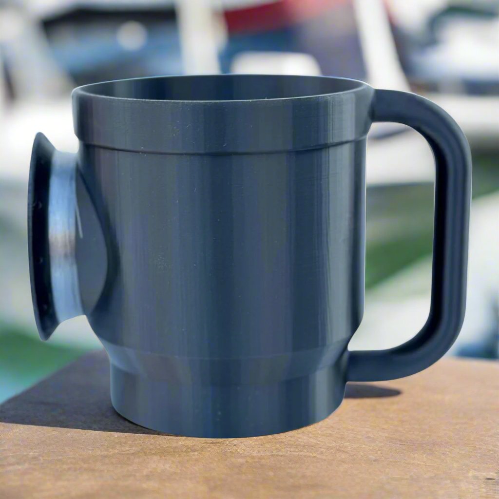 A blue mug with a large, round suction cup on its side rests on a wooden surface, evoking the design of a Chill-N-Reel® Tumbler Handle. The background is blurred, hinting at an outdoor environment with boats or water.