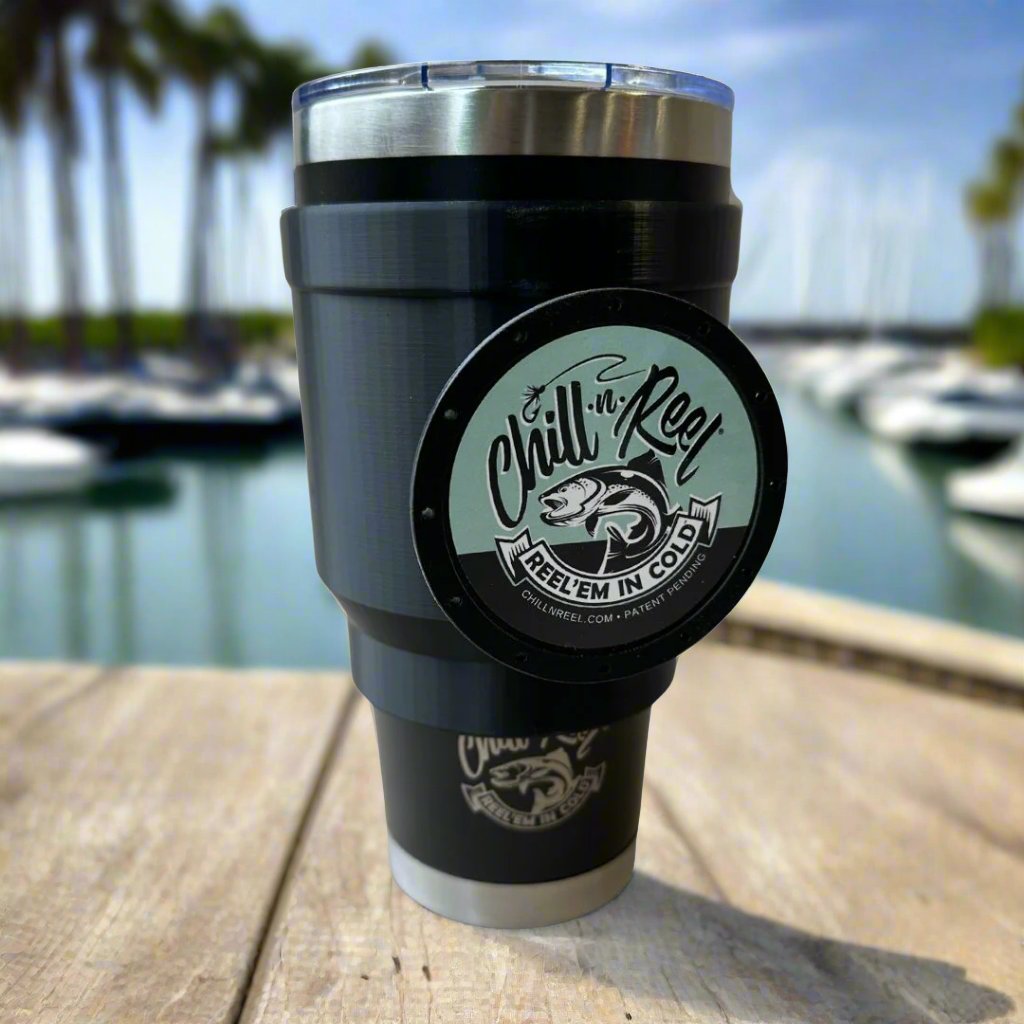 A sleek Chill-N-Reel® 30oz Tumbler topped with the innovative Chill-N-Reel Tumbler Handle (Prototype) rests on a wooden table, making for the ideal fishing companion. It overlooks a marina framed by boats and palm trees under a clear, sunny sky.