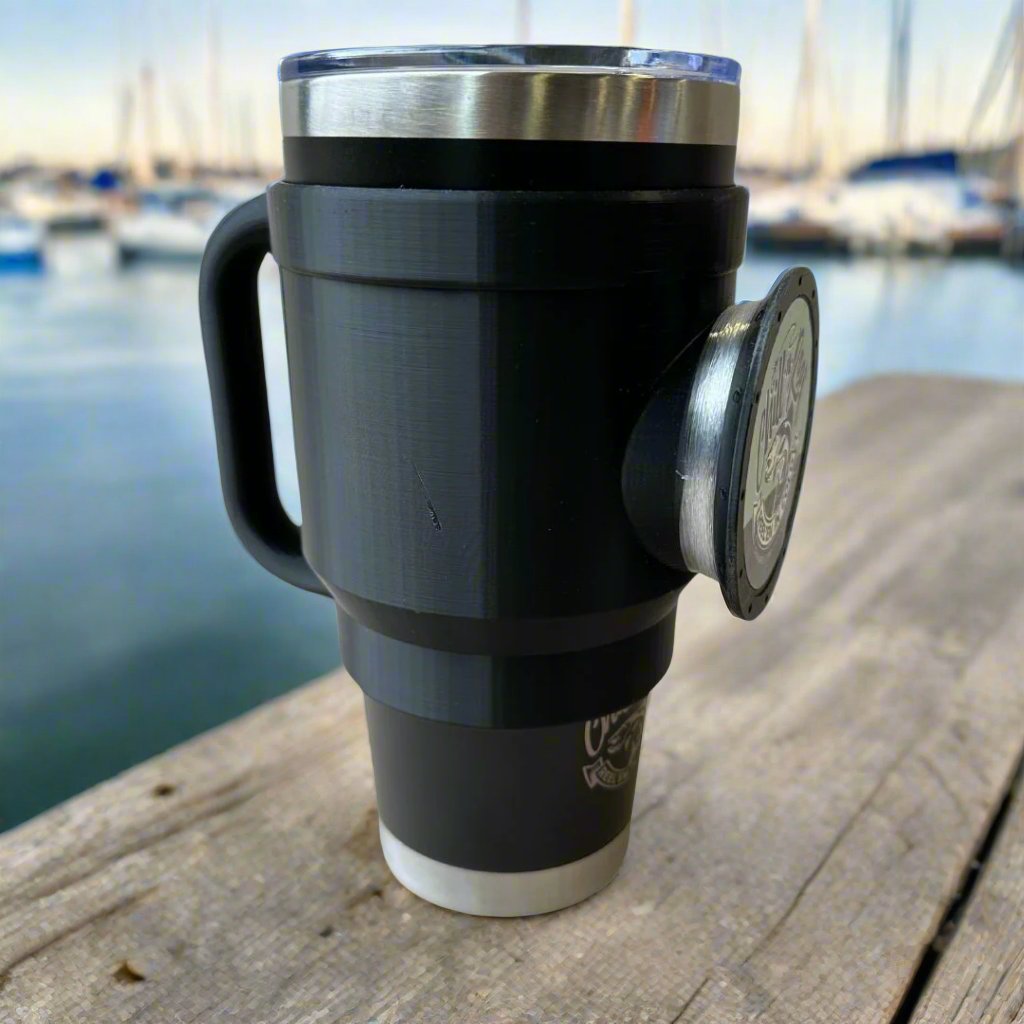 A Chill-N-Reel® Tumbler Handle Prototype with a 30oz black tumbler rests on a wooden surface, highlighting the convenience of the Chill-N-Reel Handle while offering a charming backdrop of a blurred marina, accentuating its appeal as both a functional fishing and drinking accessory.