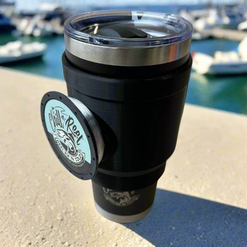 A Chill-N-Reel Tumbler Handle (Prototype) with a 30oz Tumbler, branded with the iconic circular logo and fitted with a transparent lid, rests on a concrete surface. In the background, a blurred marina featuring boats shines under the clear blue sky, perfectly capturing tranquil fishing vibes.