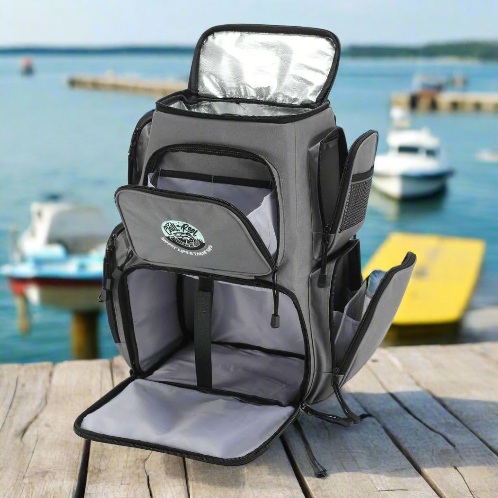 A gray fishing tackle backpack from Chill-N-Reel® features multiple compartments with zippered pockets, including a large main open section and smaller side pockets. The top flap is open, showcasing an insulated lining ideal for cooler storage. The logo on the front pocket reads "Chill-N-Reel®.