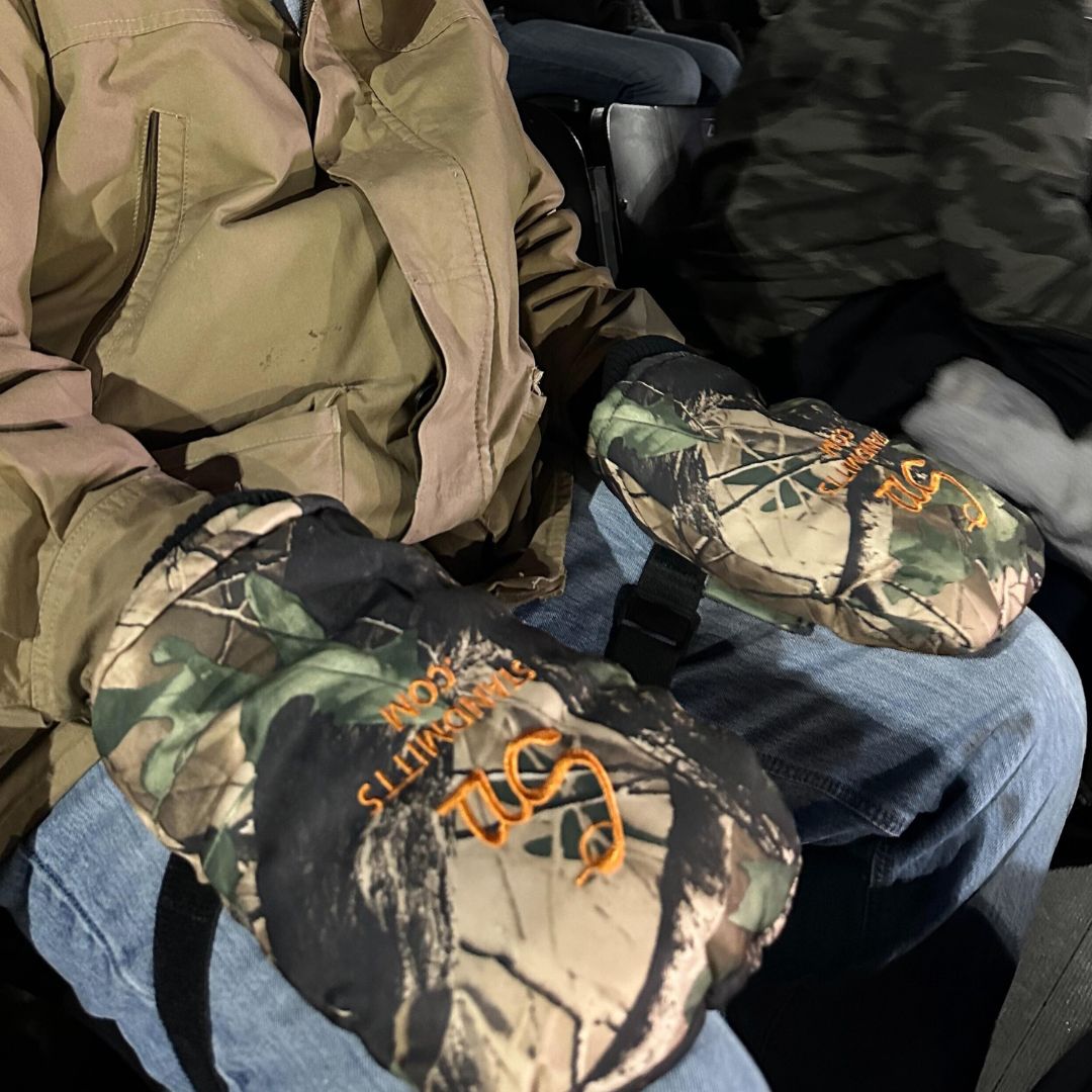A person wearing blue jeans and a beige jacket is sitting down. They have large Stand Mitts by Chill-N-Reel®: Warm Thumbless Mittens for Hunting, Ice Fishing, Sporting Events, Camping. Strap to Legs, Tree Stands, Chairs on their lap with the letters "SW" and "Smokey's Sports" embroidered in orange, ideal for winter outdoor activities.