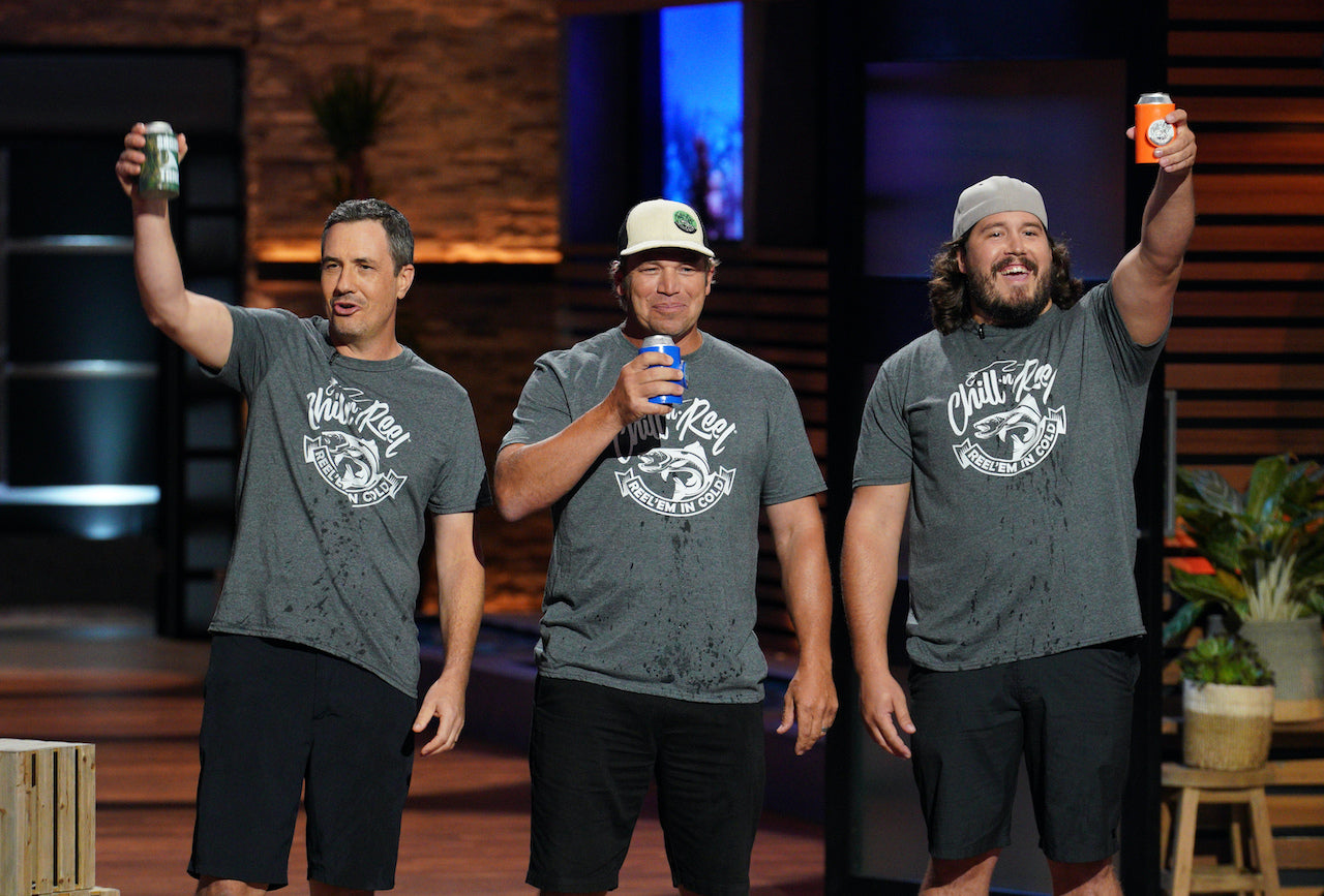 Three men stand on a stage, each holding up a can. They are wearing matching "Chill-N-Reel" t-shirts and black shorts. One man wears a white bucket hat, another a grey backwards cap. The backdrop has a brick wall on one side and dark paneling on the other.