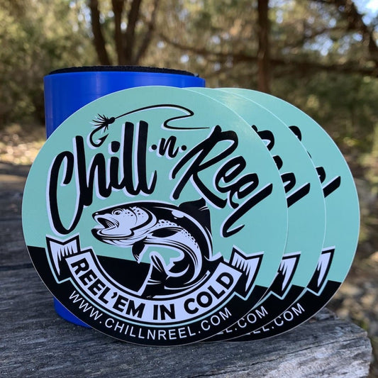 Three round "Vinyl Sticker - 4\"x4\" - Chill-N-Reel" stickers from Chill-N-Reel are placed on a wooden surface outdoors with a blue drink holder in the background. The stickers feature a design of a fish and fishing hook with the text "Chill-N-Reel" and "Reel’em in Cold" along with the website URL—perfect for your truck or boat.