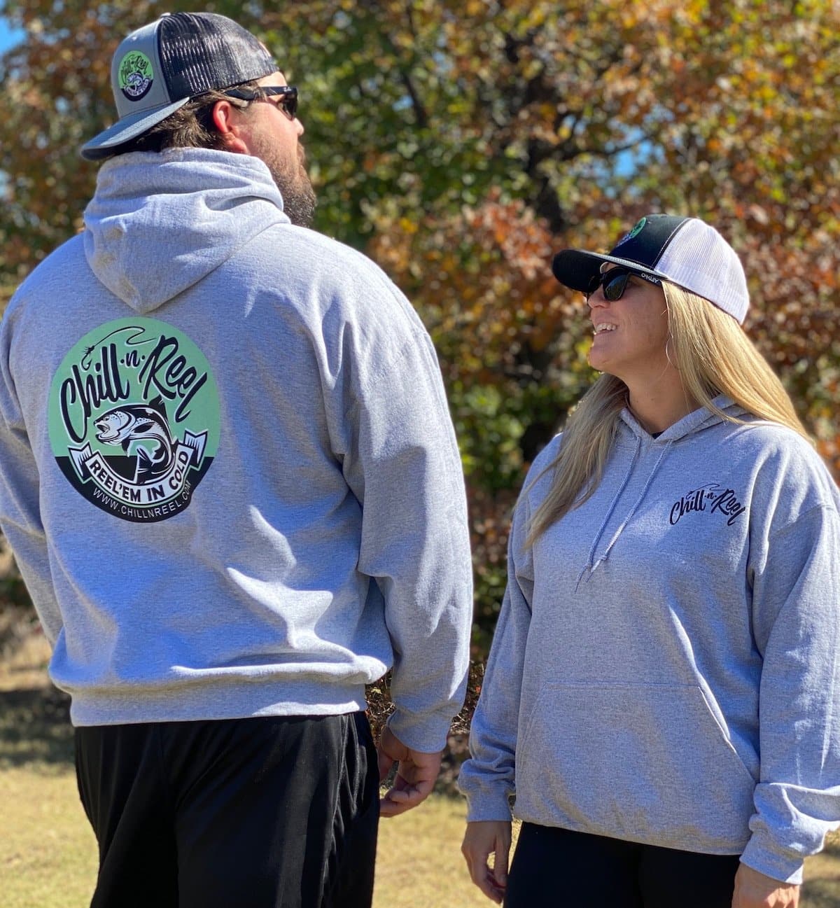 Two people are standing outdoors in Hoodie - Chill-N-Reel by Chill-n-Reel, classic athletic fit gray hoodies with the "Chill-n-Reel" text and a fish logo. They are facing each other, smiling. The man is also wearing a black cap and sunglasses, while the woman sports a black and white cap and sunglasses. Trees with autumn leaves provide a cozy backdrop.
