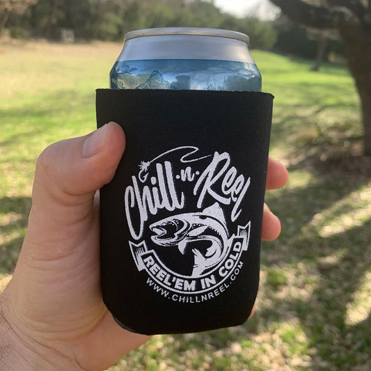 A hand holds a can wrapped in a black Chill-N-Reel Coolie Sleeves (spare inserts for Chill-N-Reel) with the text "Reel’Em In Cold" and a fish graphic. Made from durable polyurethane foam cozies, it keeps your drink cold while you fish. The background features a grassy field with trees. Visit www.chillnreel.com for more details.