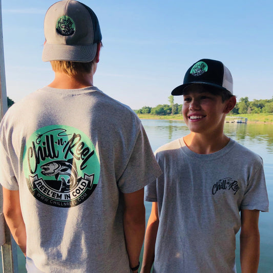 Two boys in gray short-sleeve Chill-N-Reel shirts and hats stand next to a body of water. One boy faces away, showing the "DryBlend Short Sleeve T-shirt - Chill-N-Reel CLASSIC COLOR LOGO (on BACK)" on his Chill-n-Reel t-shirt, while the other smiles at the camera. Trees and a dock are visible in the background.
