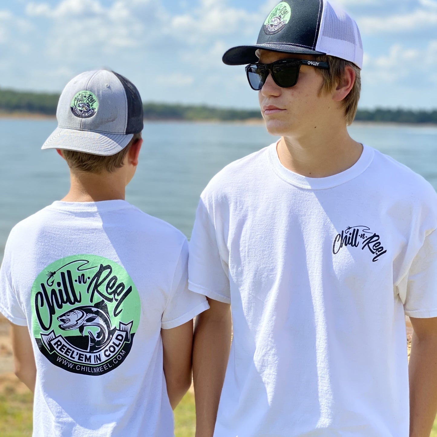 Two people are standing by a body of water. Both are wearing white Chill-n-Reel DryBlend Short Sleeve T-shirt - Chill-N-Reel CLASSIC COLOR LOGO (on BACK), baseball caps, and sunglasses. One person faces the camera, while the other is turned away, showing the back of their t-shirt which features a large logo with a fish.