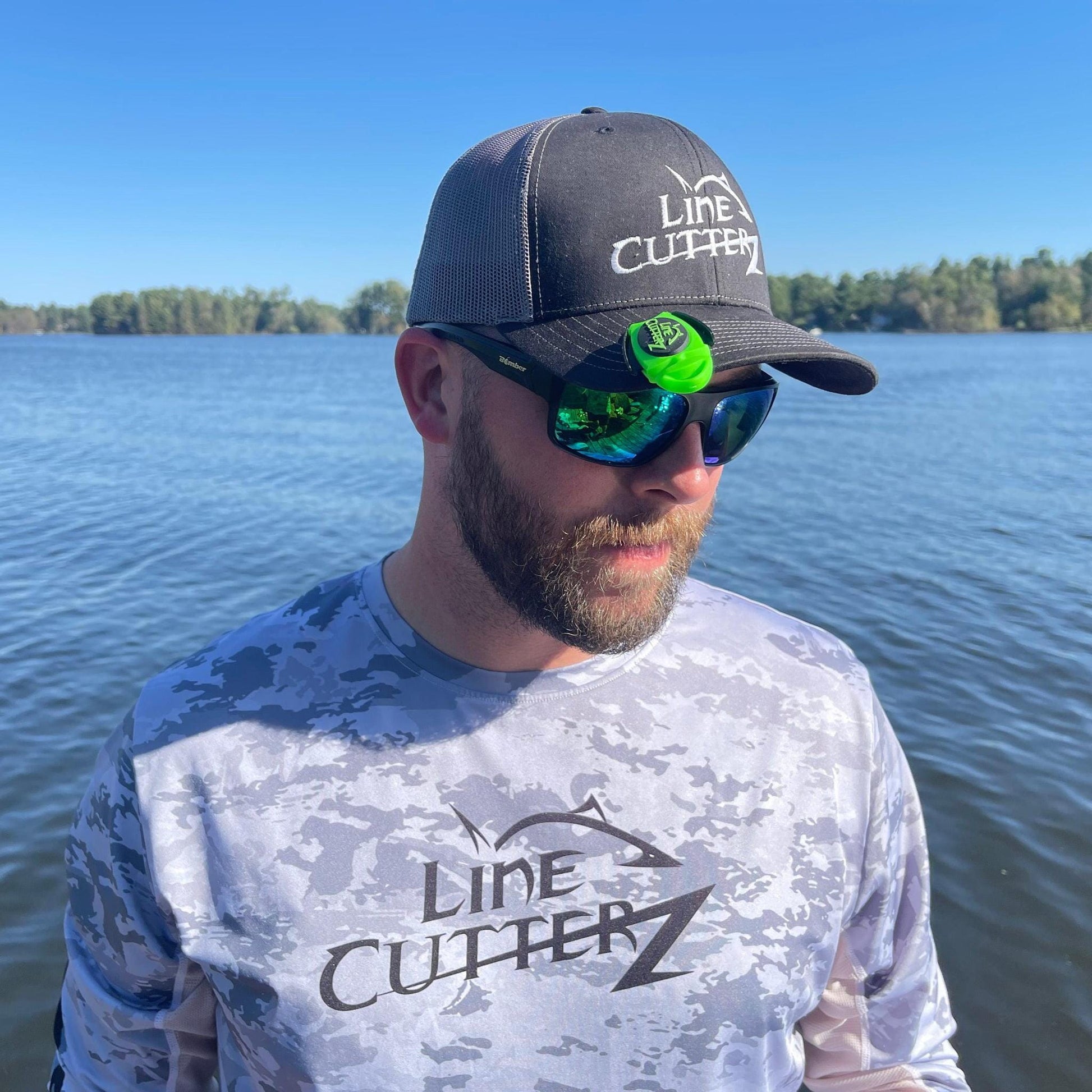 A bearded man in a Line Cutterz hat and shirt stands lakeside, wearing reflective sunglasses with a Line Cutterz Dual Hybrid Micro Scissors attached—highlighting their innovative ceramic cutting technology against a scenic backdrop of trees and blue sky.