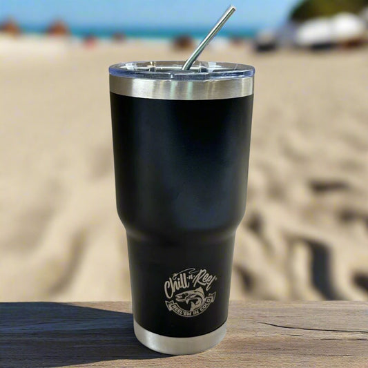 A 30oz Stainless Steel Vacuum Insulated Tumbler by Chill-N-Reel® rests on a wooden surface against a blurred beach backdrop. It showcases the Chill-N-Reel® logo and includes a sturdy stainless steel straw, providing stylish refreshment for your seaside adventures.