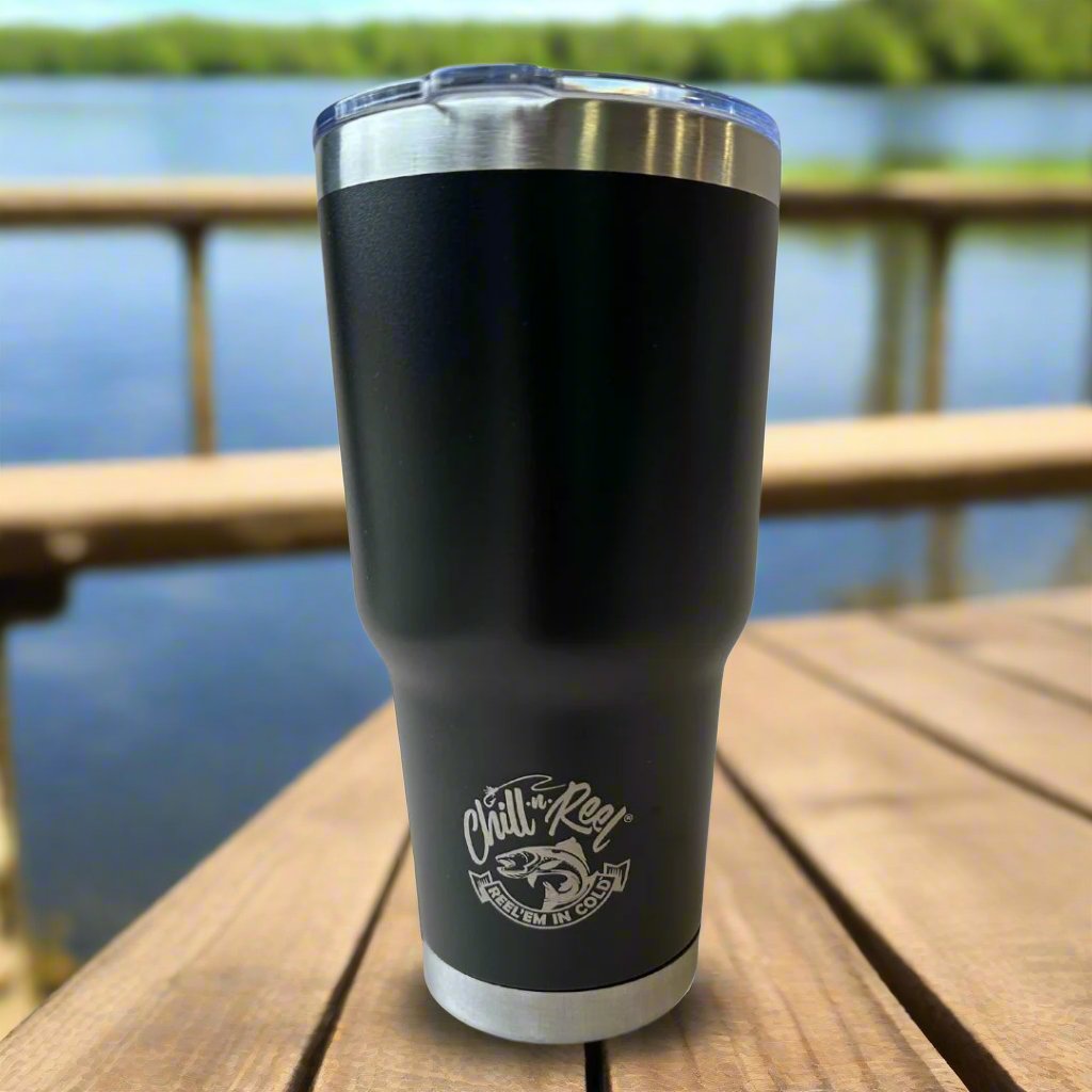 A 30oz Stainless Steel Vacuum Insulated Tumbler by Chill-N-Reel® rests on a wooden table near a lake. This tumbler, with a metal rim and clear lid, features double-wall vacuum insulation and displays a fishing-themed logo along with the phrase "Chill-N-Reel." The backdrop offers a blurred glimpse of water and lush green trees.