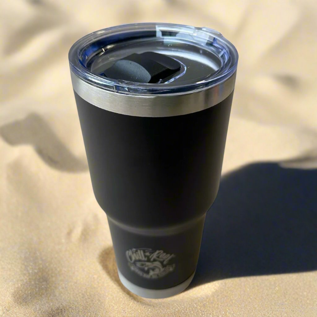 A 30oz Stainless Steel Vacuum Insulated Tumbler by Chill-N-Reel® is set on the sandy ground. It features a clear lid and a logo on its lower part, casting a shadow on the sand.