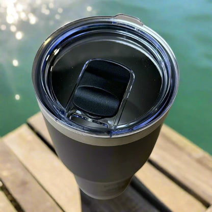 A 30oz Stainless Steel Vacuum Insulated Tumbler by Chill-N-Reel® with double-wall vacuum insulation and a transparent lid sits on a wooden surface near a shimmering body of water. Sunlight reflects off the water, creating a dazzling backdrop while ensuring your drink maintains its perfect temperature.