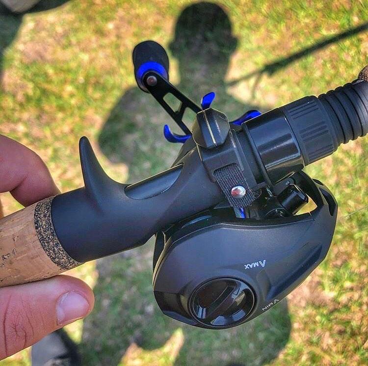 A close-up of a person holding a fishing rod with a black baitcasting reel and wearing the Line Cutterz "TRIPLE PLAY" Fishing Line Cutter Multi-Pack ring, standing on grass with visible shadows in the background.