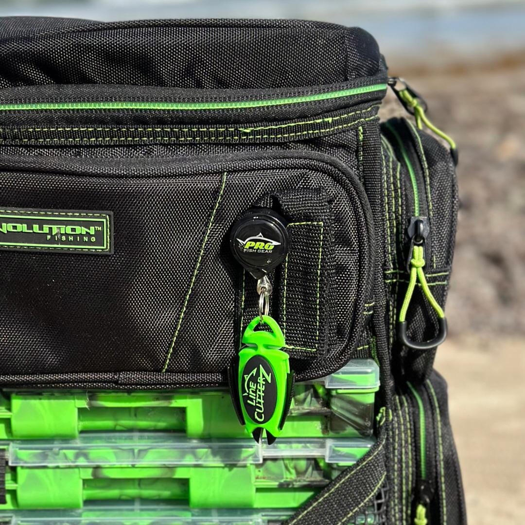 A close-up of a black and green fishing tackle bag with a "Pro" branded retractable clip and Line Cutterz Dual Hybrid Micro Scissors attached to the front zipper pocket. Several plastic tackle boxes are visible inside the bag.