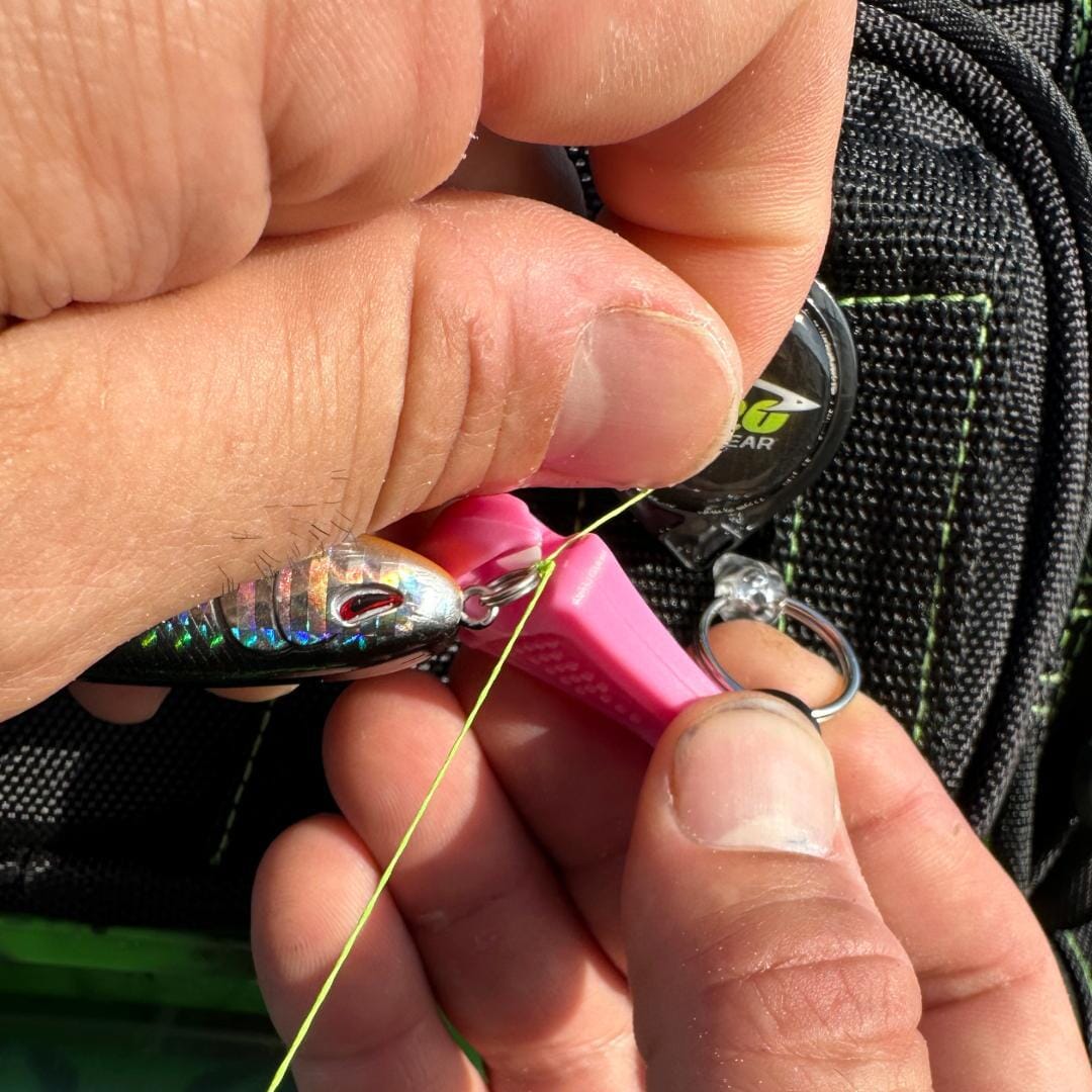 Close-up of hands threading fishing line through a fish-shaped lure, with a pink tool and a Line Cutterz Ceramic Blade Zipper Pull by Line Cutterz attached to a black bag with green stitching in the background.