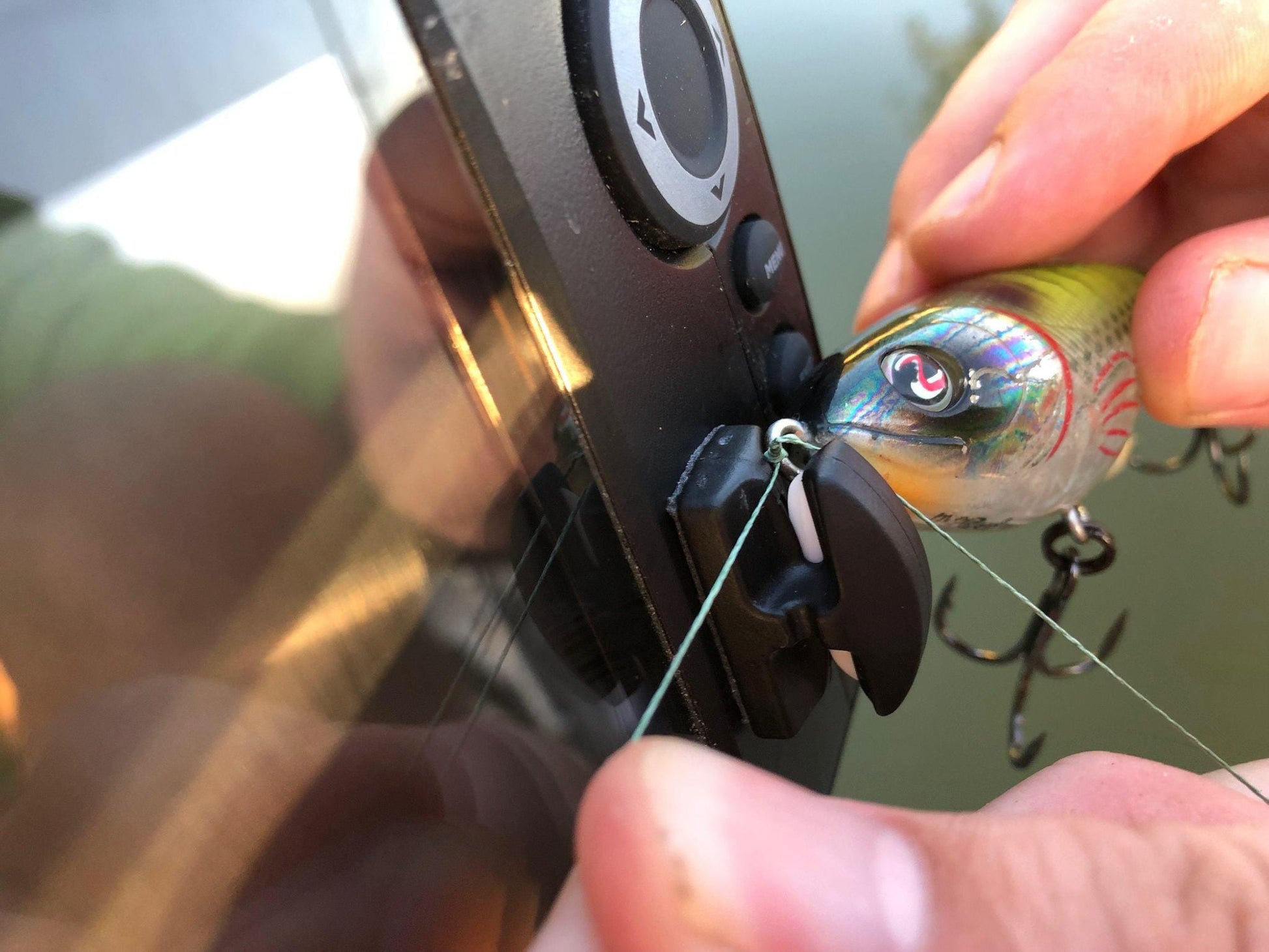 Close-up of hands tying fishing line with a lure next to a Line Cutterz Ceramic Blade Peel & Stick Flat Mount, a rust-proof line cutter by Line Cutterz, all illuminated by outdoor sunlight.