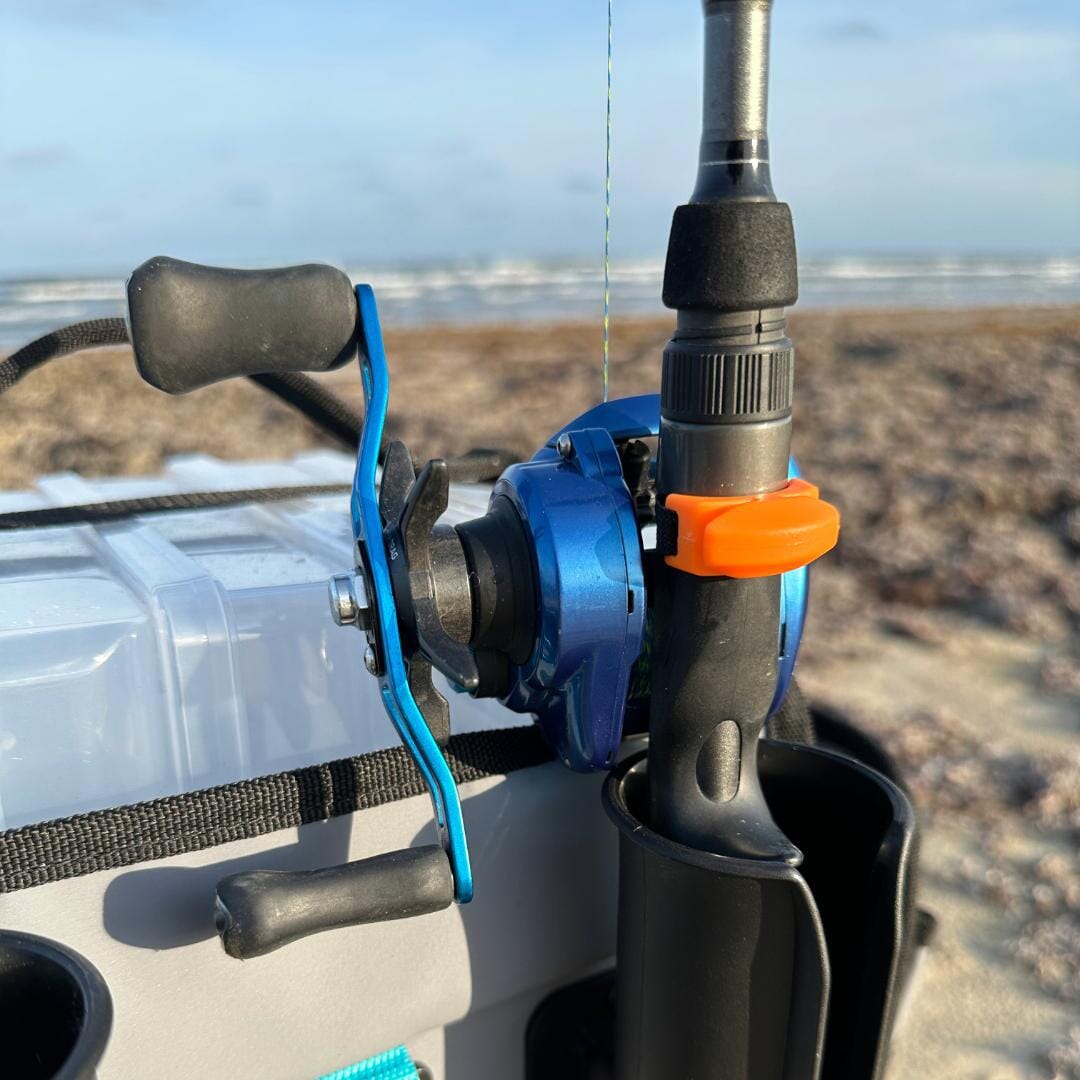 Close-up of a blue fishing reel with a Line Cutterz Ceramic Blade Ring by Line Cutterz attached to a black rod; tackle box and sandy beach behind, with ocean and sky visible in the distance.