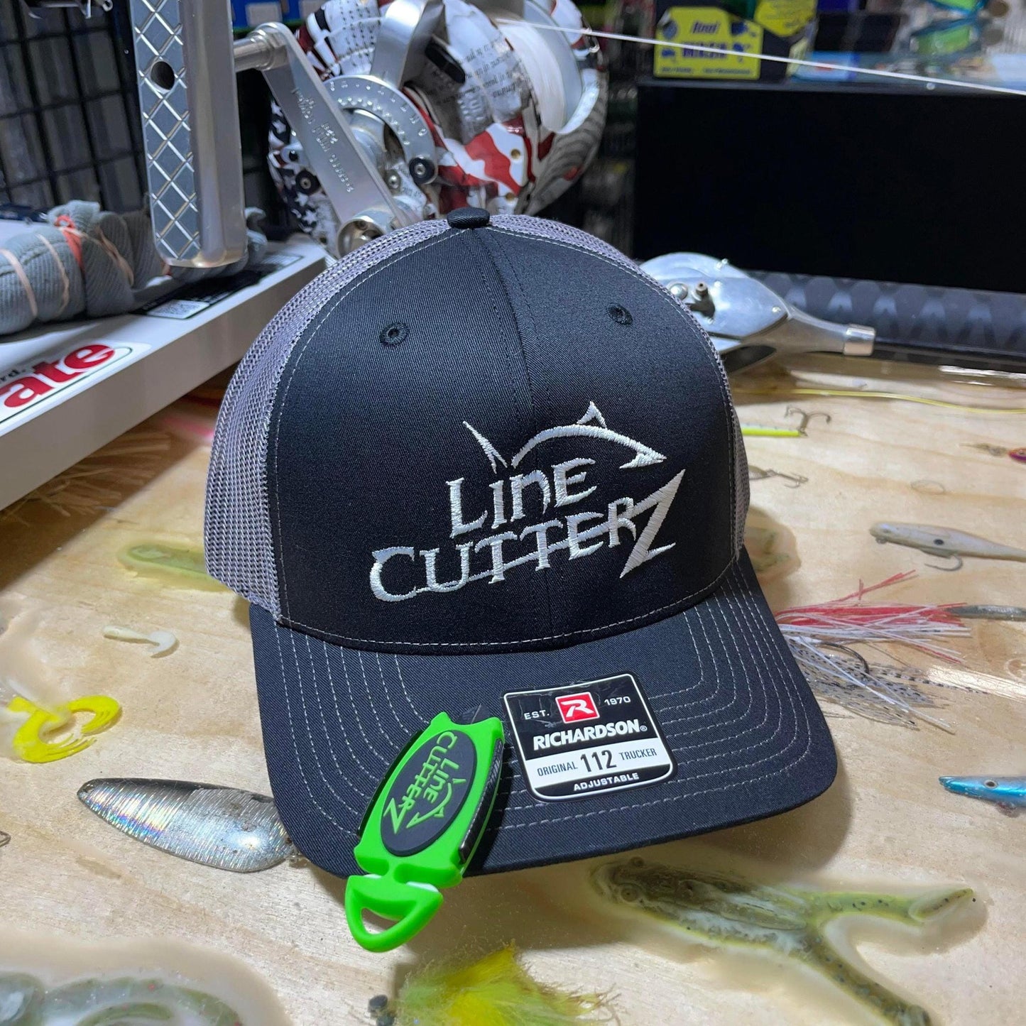 A black and gray mesh-back cap with "Line Cutterz" embroidered on the front sits among fishing gear, with a green Line Cutterz Dual Hybrid Micro Scissors featuring ceramic cutting technology attached to the bill.