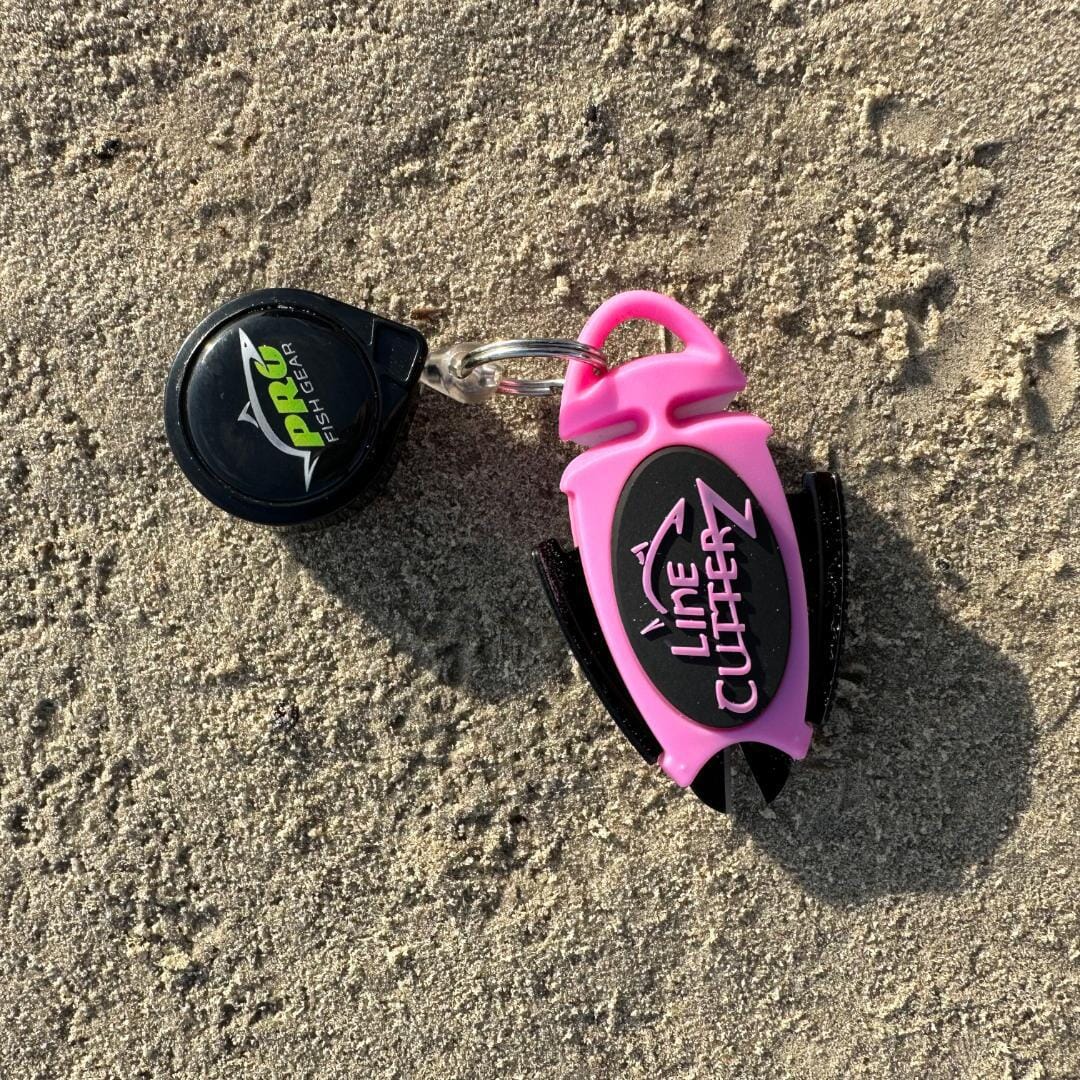 A pink Line Cutterz Dual Hybrid Micro Scissors with ceramic blades and a black ProFish gear retractable clip, connected by a keyring, lie on sandy ground.