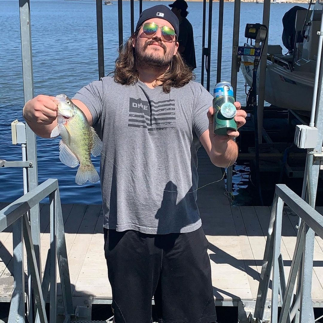 A man with long hair, wearing sunglasses and a cap backward, stands on a dock holding a fish in one hand and a can in the other. He is wearing a grey T-shirt and black shorts. A body of water and a boat are visible in the background. The weather is clear and sunny.