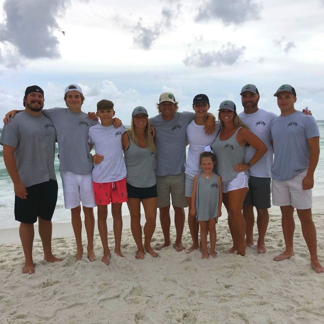 A group of nine adults and one child standing close together on a sandy beach. They are all wearing matching grey shirts and hats. The adults are all casually dressed in shorts and T-shirts, while the child is in a tank top and shorts. The ocean and cloudy sky are in the background.