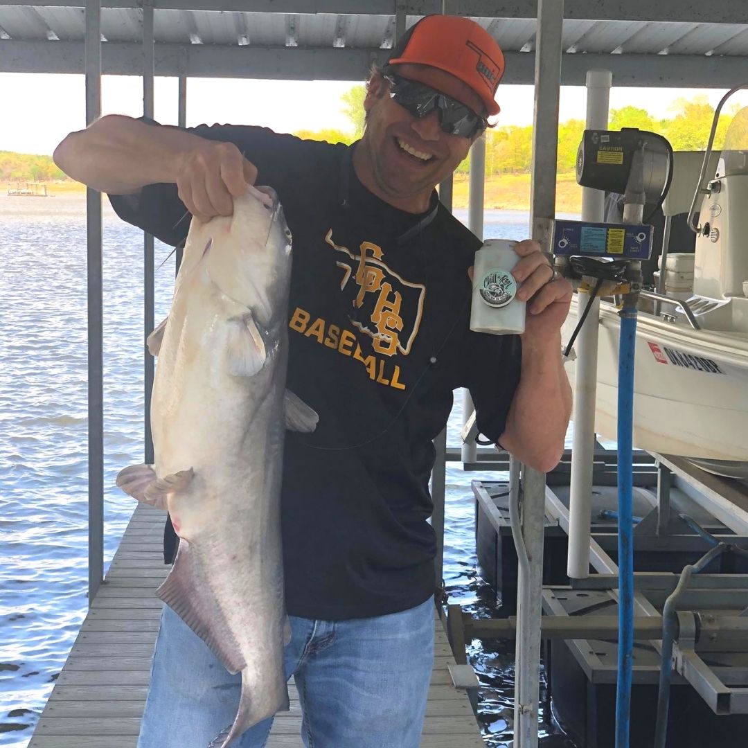 A person stands on a dock holding up a large fish with one hand and a canned beverage in the other. They wear sunglasses, a black "Baseball" T-shirt, jeans, and an orange cap. A boat and waterway are visible in the background. The person is smiling.