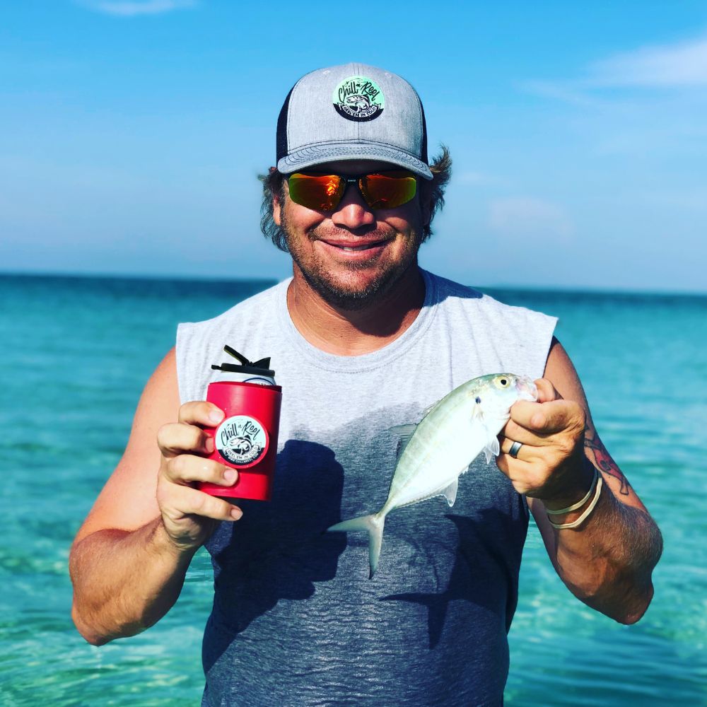 A person wearing a grey sleeveless shirt, sunglasses, and a hat holds a small fish in one hand and a red beverage can with a "Wild Bills" logo in the other hand. They are standing in shallow, clear blue water under a sunny sky.
