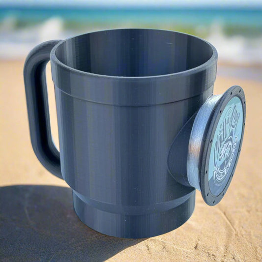 A Chill-N-Reel® Tumbler Handle in a large gray 30oz size rests on a sandy beach, featuring an attached lid with a wave design and text that add flair. The blurred backdrop of the beach and ocean waves creates the perfect setting for relaxation after enjoying a fishing game.