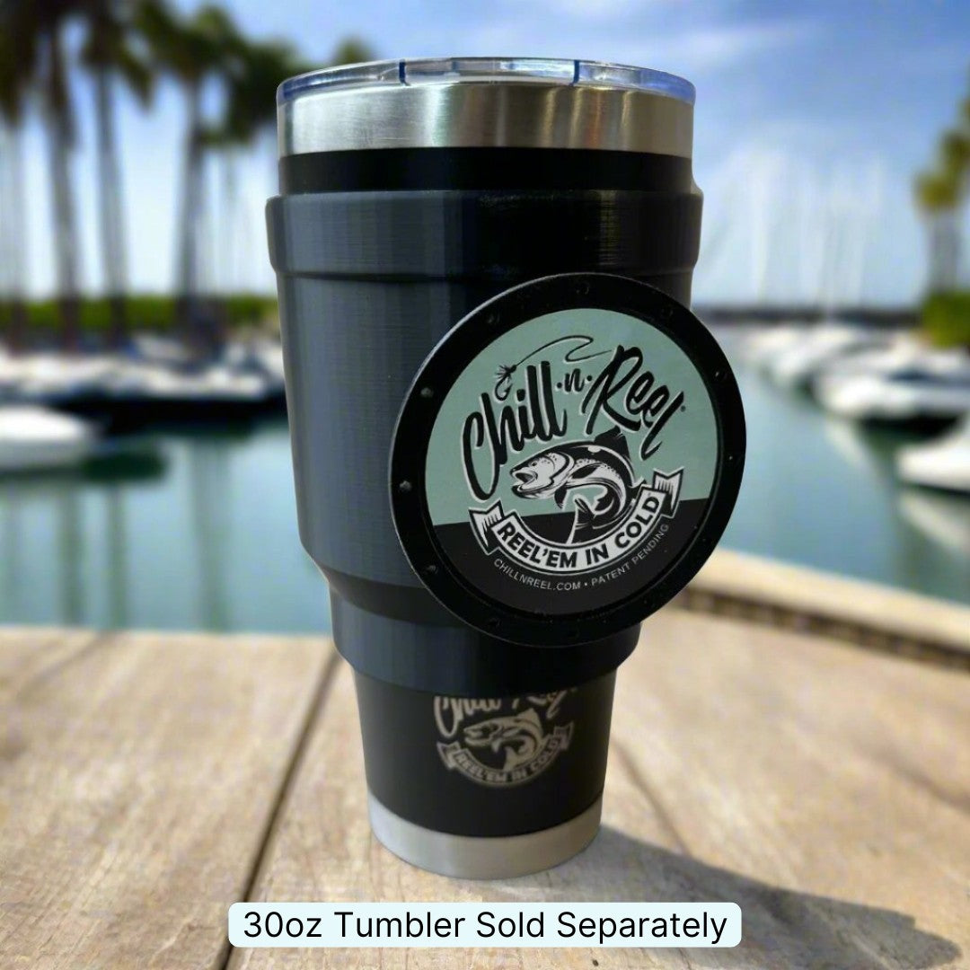 A black 30oz tumbler with the Chill-N-Reel® logo and a fish graphic is set on a wooden surface. In the backdrop, blurred boats and palm trees at a marina provide the scenery under a clear sky. This tumbler handle is ideal for fans of the popular fishing game, though it must be purchased separately.