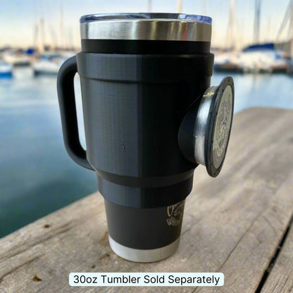 A black tumbler with a 30oz capacity, showcasing the Chill-N-Reel® Tumbler Handle and a removable metal badge, is placed on a wooden surface. The background features a marina with boats slightly out of focus. A caption underneath states, "30oz Tumbler Sold Separately." Ideal for those passionate about fishing.