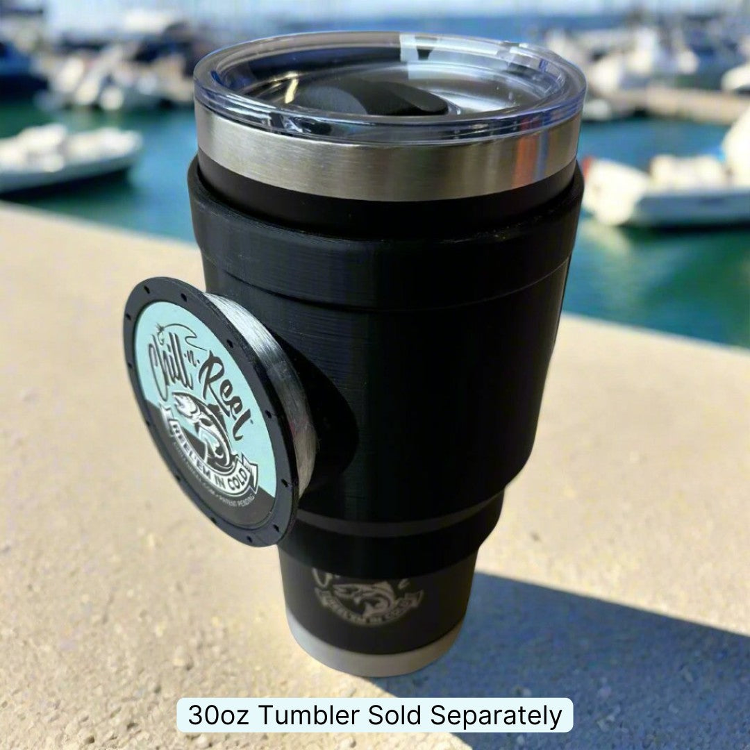 A black 30oz Chill-N-Reel® Tumbler Handle with a clear lid sits on a ledge by the marina, boats gently rocking in the background. Featuring a circular logo on its side, this limited prototype radiates charm. Caption reads, "30oz Tumbler Sold Separately.