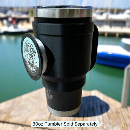 A black 30oz tumbler equipped with a Chill-N-Reel® Tumbler Handle rests on a wooden surface by the marina. The background showcases blurred boats and water, capturing the essence of a tranquil fishing game scene. The text states "30oz Tumbler Sold Separately.