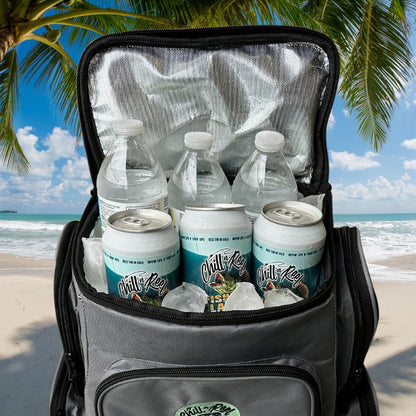 A Chill-N-Reel® Fishing Tackle Backpack with Cooler Top, filled with ice-cold water bottles and canned drinks, is open on a sandy beach beneath the shade of palm trees, set against a clear blue sky.