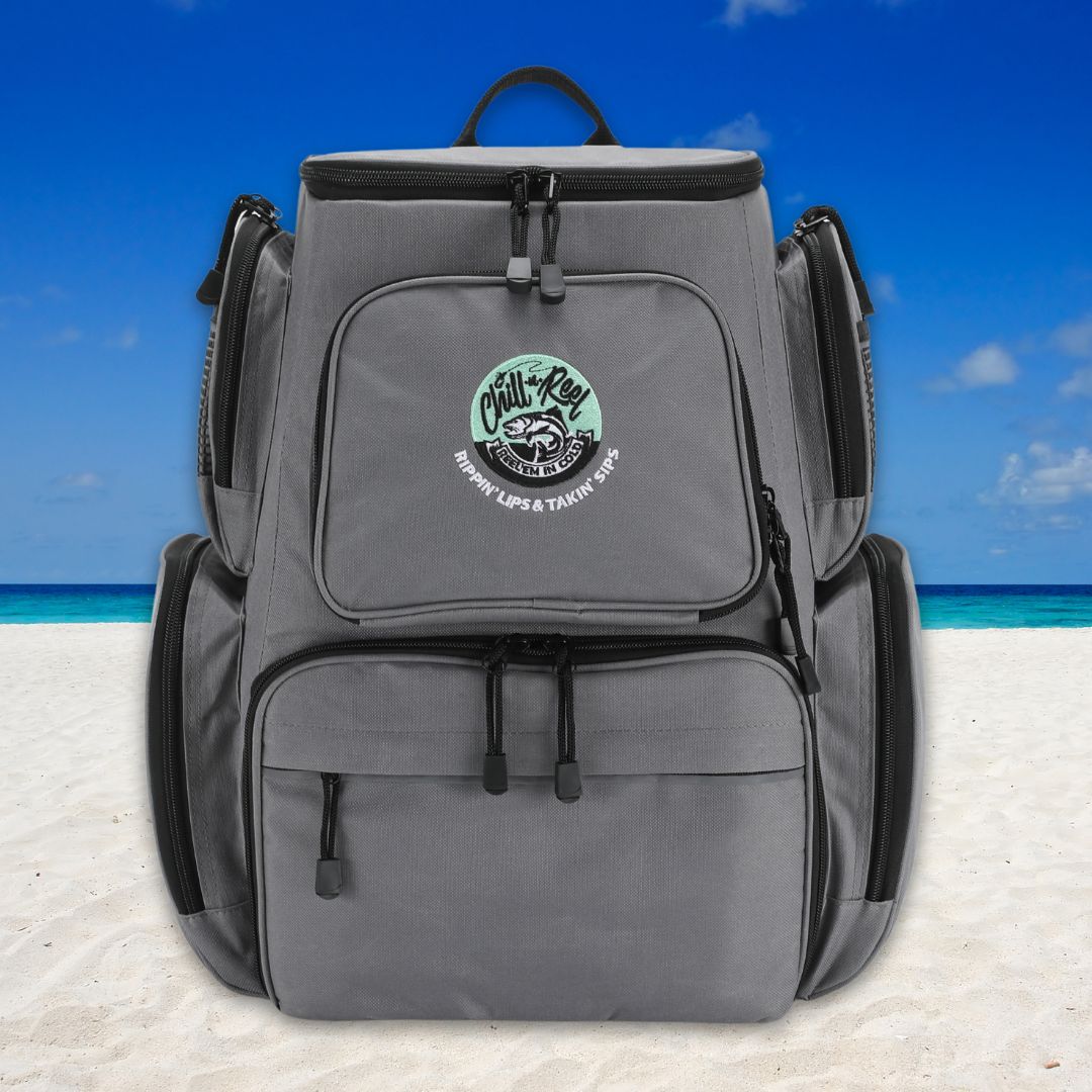 A Fishing Tackle Backpack with Cooler Top by Chill-N-Reel®, featuring a gray design with multiple compartments and black zippers, sits on a sandy beach under a clear blue sky. A patch on the front displays "Chill-N-Reel®" within a circular design, adding an adventurous touch.