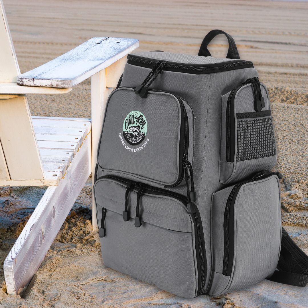 A Chill-N-Reel® Fishing Tackle Backpack with Cooler Top, boasting multiple compartments, rests on a sandy beach beside a white wooden chair. This gray backpack, adorned with the Chill-N-Reel logo and text, is designed for optimal tackle storage and includes a convenient cooler top.