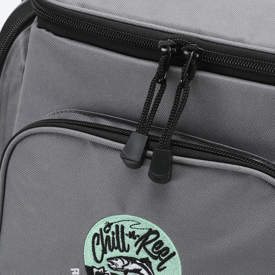 Close-up of the Chill-N-Reel® Fishing Tackle Backpack with Cooler Top, featuring black zippers. The front pocket showcases an embroidered patch with a fish and the words "Chill Reel" in stylized lettering, ideal for those who appreciate efficient tackle storage on their fishing adventures.