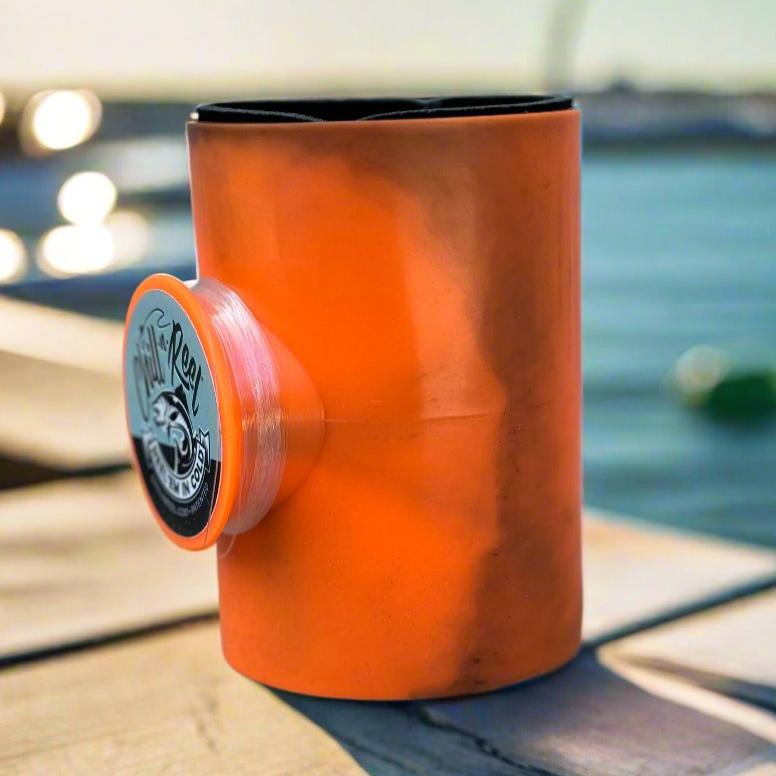 A limited edition, orange, cylindrical Mystery Color Chill-N-Reel by Chill-N-Reel® is showcased on a wooden surface near a body of water. The holder features a logo depicting a fish and accompanying text, with sunlight reflecting off the water in the background.