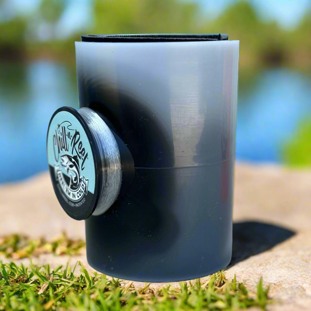 Close-up of a cylindrical Mystery Color Chill-N-Reel by Chill-N-Reel®, used for fishing. It is black and partially transparent, with a spool of fishing line attached to it. The background includes a blurred view of greenery and a body of water, indicating an outdoor setting.