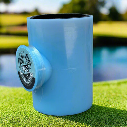 A blue, cylindrical container with an attached side holder displaying the Chill-N-Reel® logo, which features a fish and the slogan "Made in the USA." The Mystery Color Chill-N-Reel is situated on a grassy surface with a blurred water and landscape background in daylight.